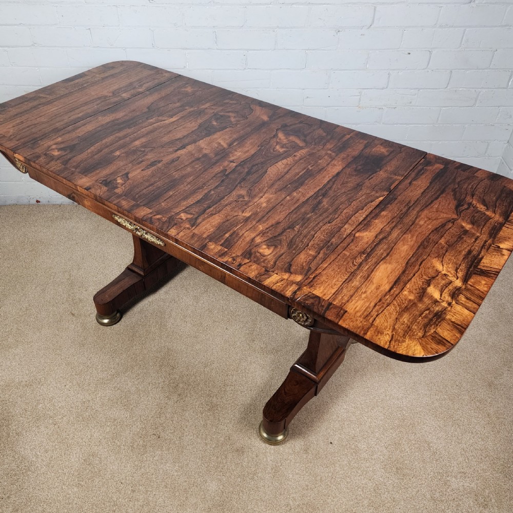 An early 19th-century Regency period rosewood sofa table featuring drop-leaf ends, twin trestle supports, decorative brass mounts, and brass castors. View IX
