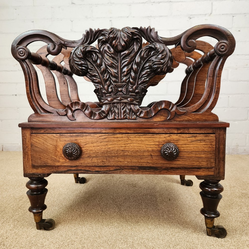 An early 19th-century carved rosewood Canterbury featuring vertical slatted partitions decorated with Prince of Wales feathers above a single base drawer with turned legs and brass castors. View VI