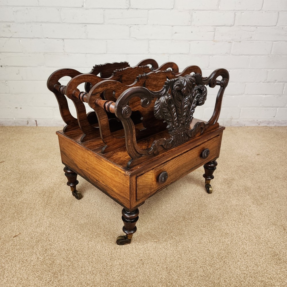 An early 19th-century carved rosewood Canterbury featuring vertical slatted partitions decorated with Prince of Wales feathers above a single base drawer with turned legs and brass castors. View VII