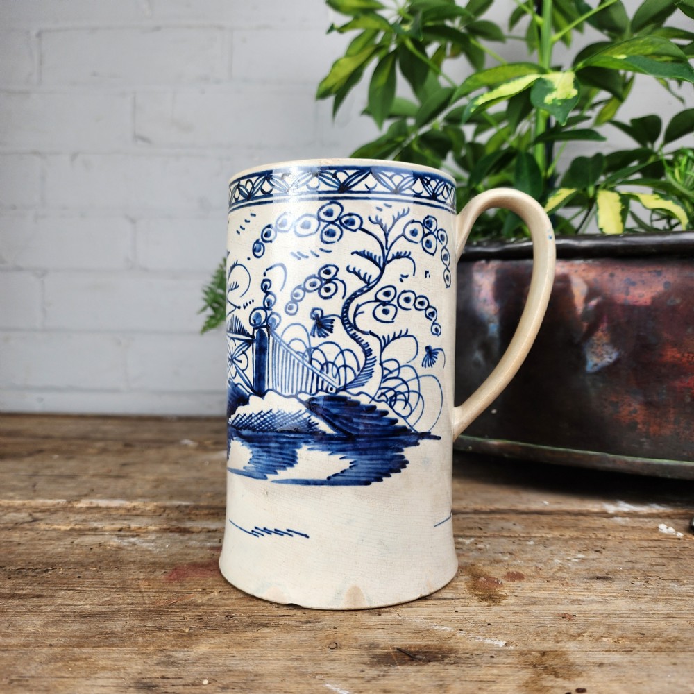 An antique cylindrical ceramic mug featuring blue and white hand-painted chinoiserie landscape decoration and an applied handle.