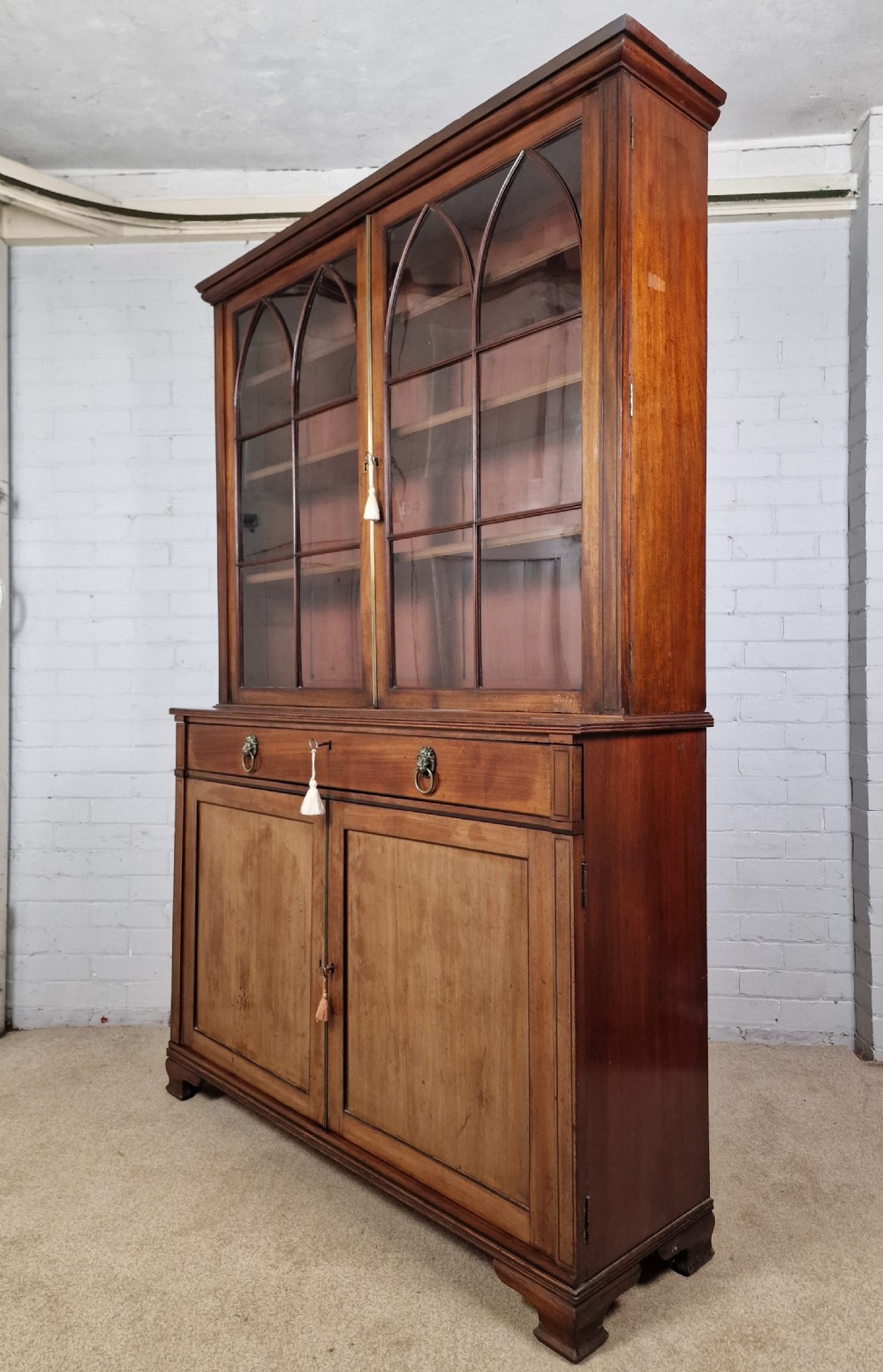 A George IV mahogany bookcase cupboard featuring glazed upper doors with Gothic arch glazing bars above a lower panelled cupboard. View XII
