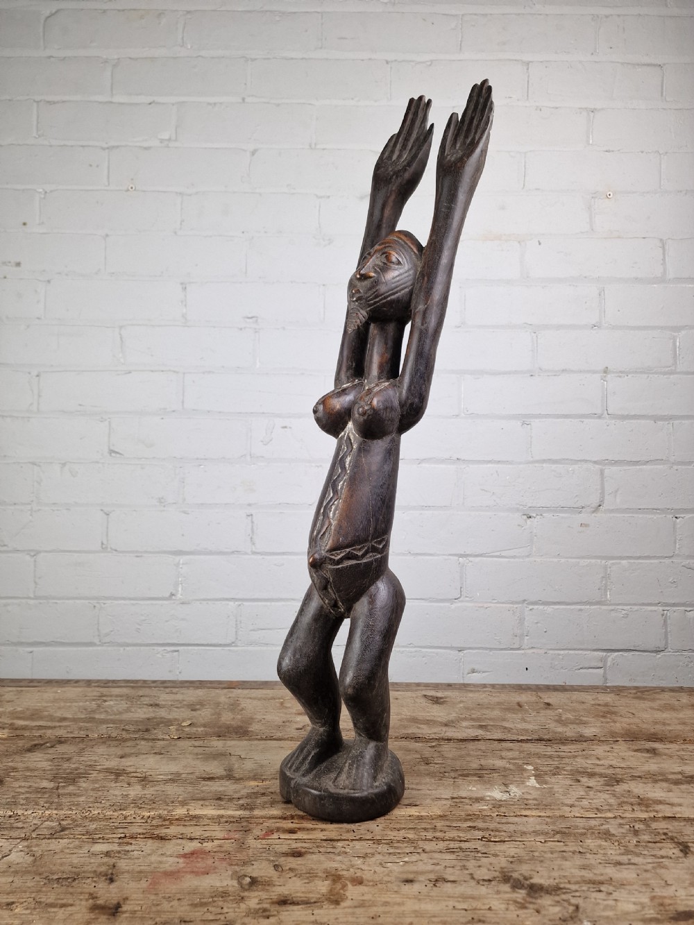 A dark carved hardwood figure depicting a female standing with both arms raised upwards above her head, mounted on an integrated circular base.
