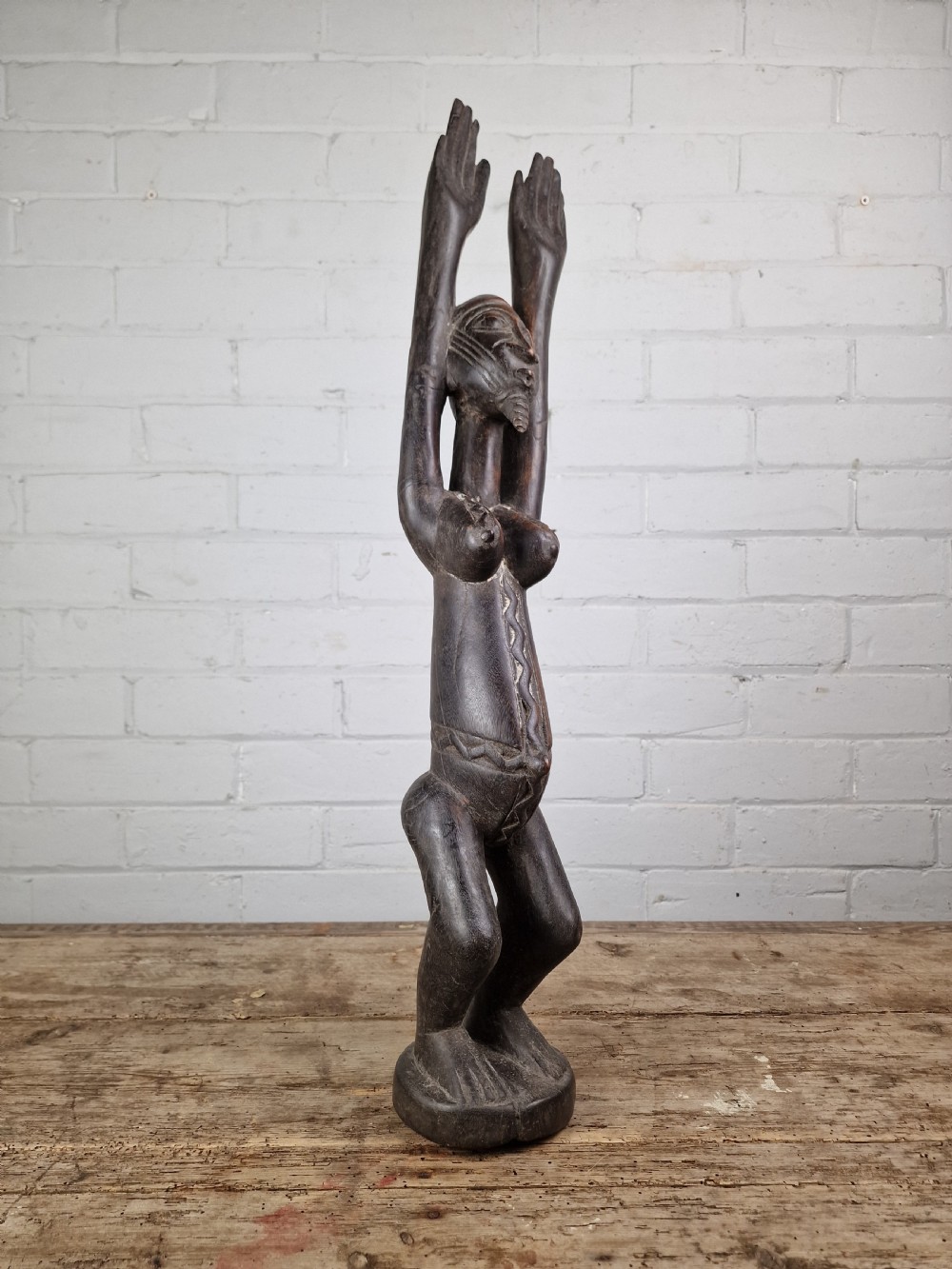 A dark carved hardwood figure depicting a female standing with both arms raised upwards above her head, mounted on an integrated circular base. View II