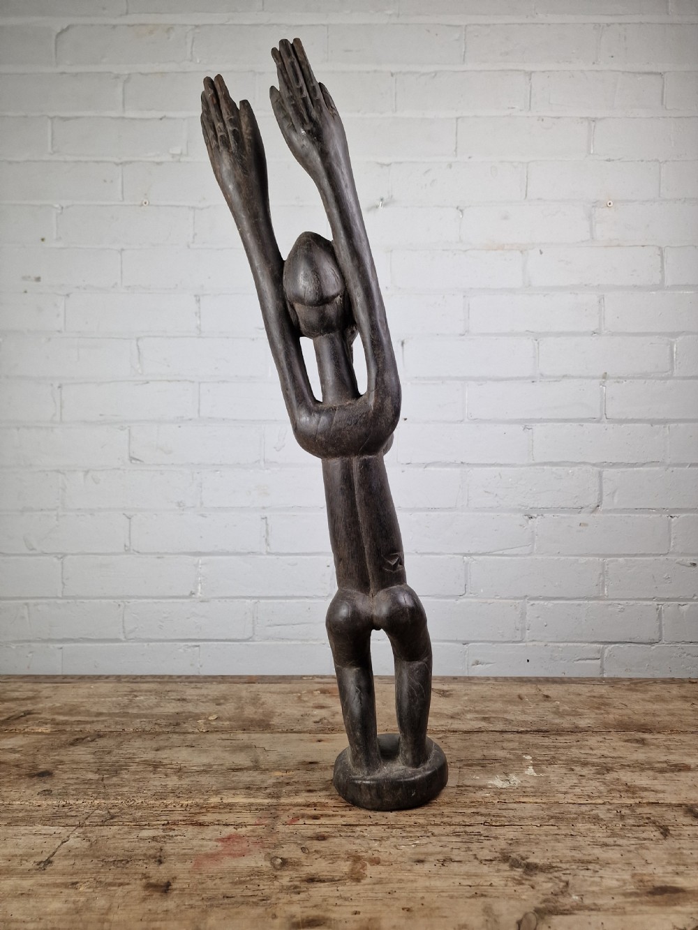 A dark carved hardwood figure depicting a female standing with both arms raised upwards above her head, mounted on an integrated circular base. View III