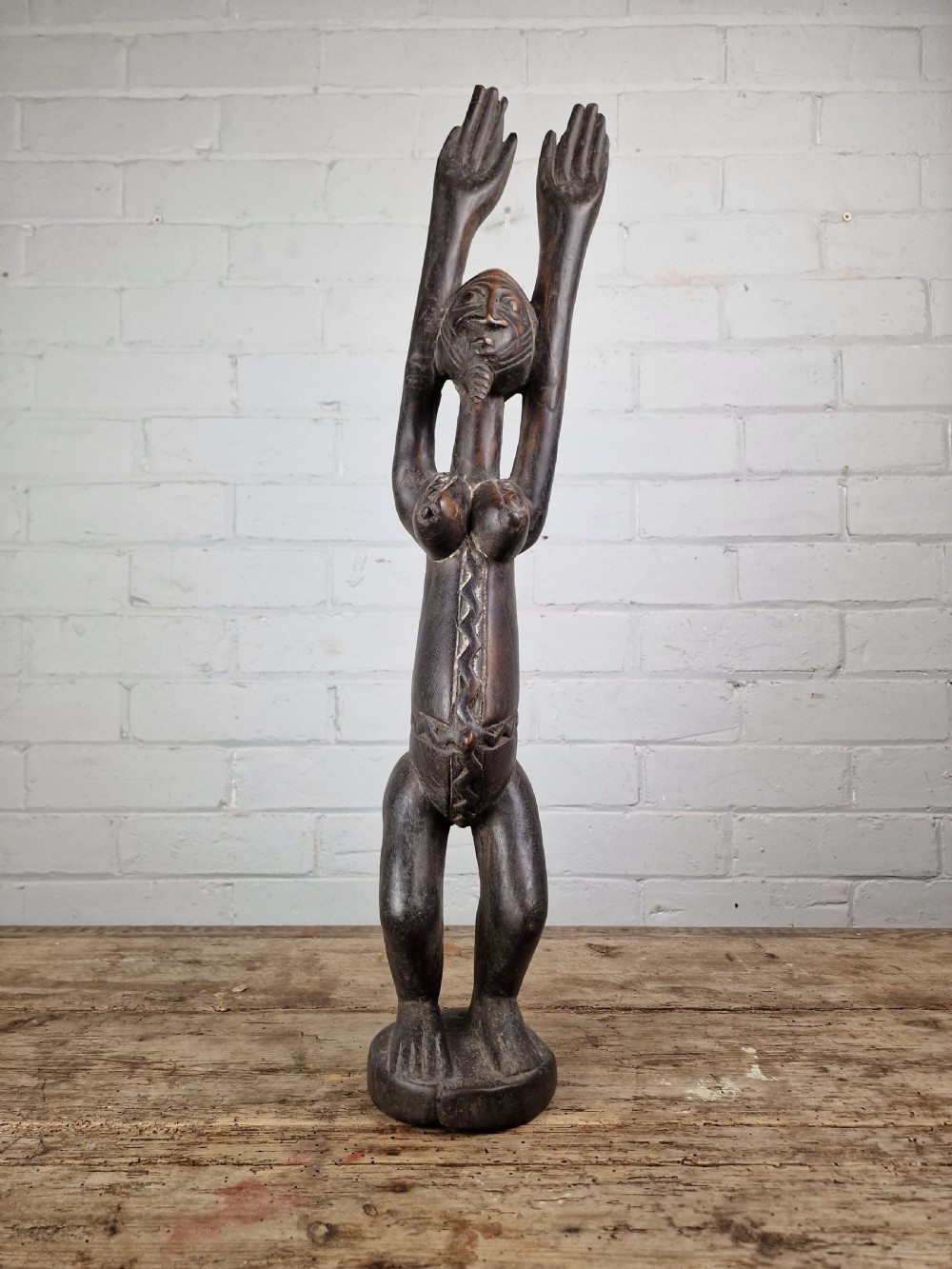 A dark carved hardwood figure depicting a female standing with both arms raised upwards above her head, mounted on an integrated circular base. View IV