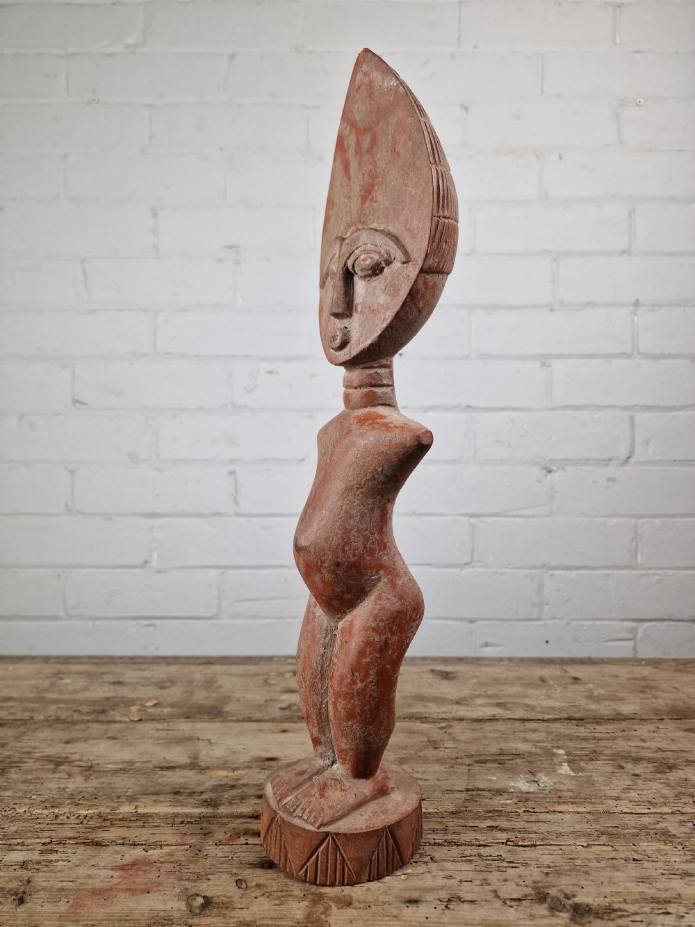 A hand-carved wooden standing Fante figure featuring an elongated crescent head, ringed neck, and a carved circular base in a reddish-brown colour.