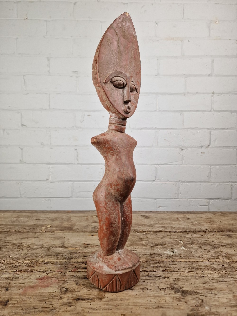 A hand-carved wooden standing Fante figure featuring an elongated crescent head, ringed neck, and a carved circular base in a reddish-brown colour. View III