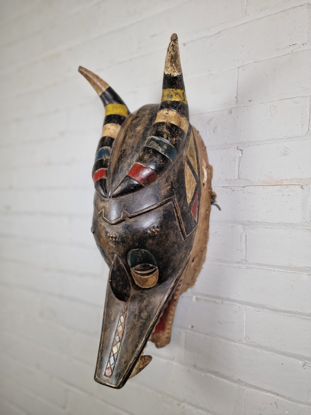 A carved wooden Guro Zamble helmet mask featuring an open-jawed snout with carved white teeth, red painted interior, dark horns, and woven fibre stitching along the base. View II