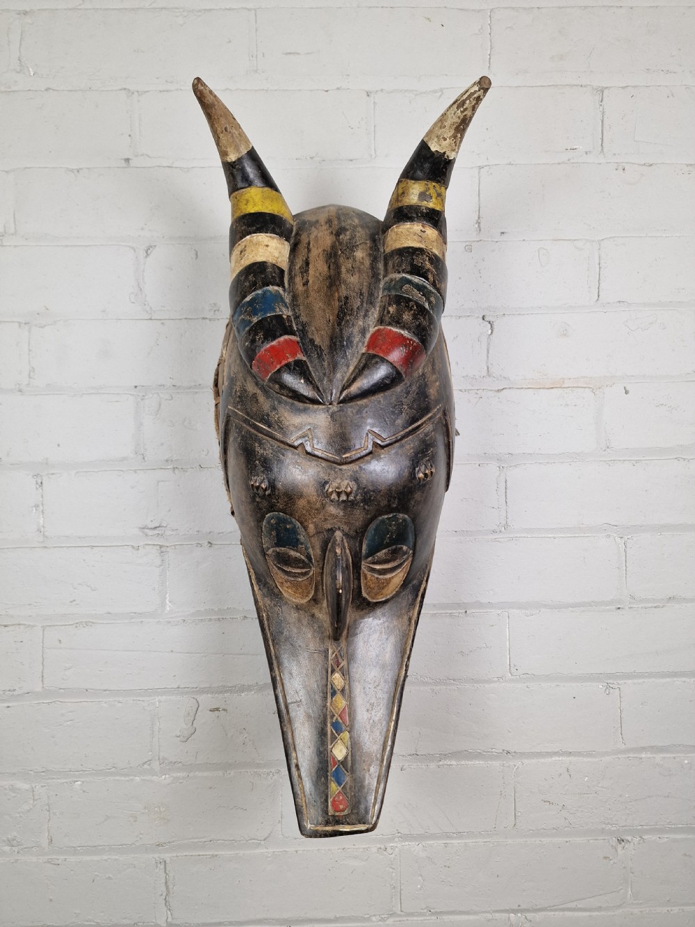 A carved wooden Guro Zamble helmet mask featuring an open-jawed snout with carved white teeth, red painted interior, dark horns, and woven fibre stitching along the base. View III