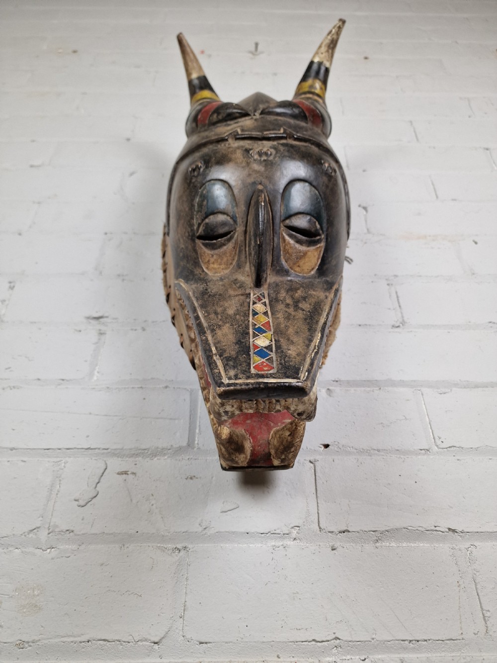 A carved wooden Guro Zamble helmet mask featuring an open-jawed snout with carved white teeth, red painted interior, dark horns, and woven fibre stitching along the base. View IV