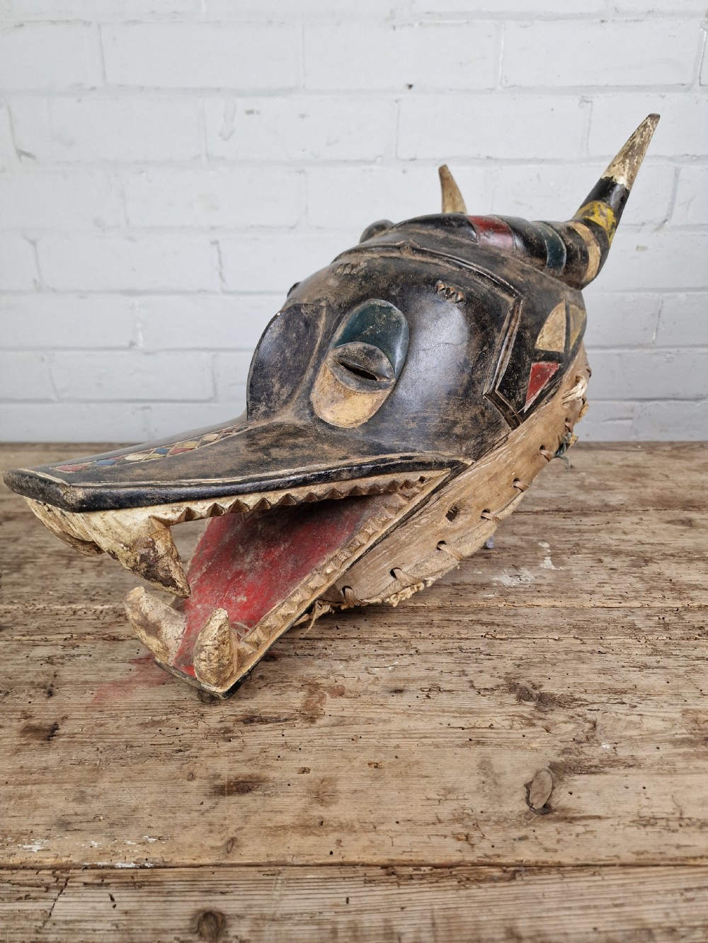 A carved wooden Guro Zamble helmet mask featuring an open-jawed snout with carved white teeth, red painted interior, dark horns, and woven fibre stitching along the base. View V