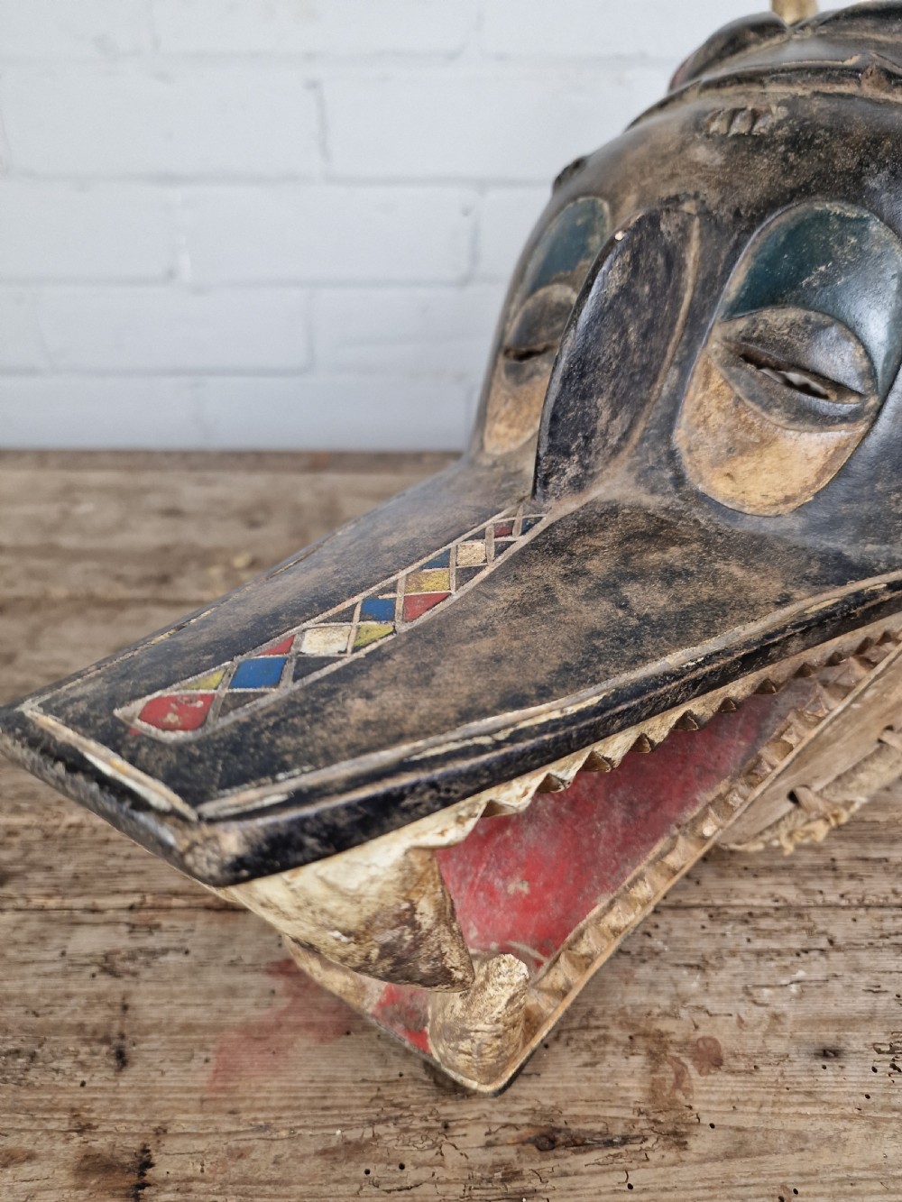 A carved wooden Guro Zamble helmet mask featuring an open-jawed snout with carved white teeth, red painted interior, dark horns, and woven fibre stitching along the base. View VI