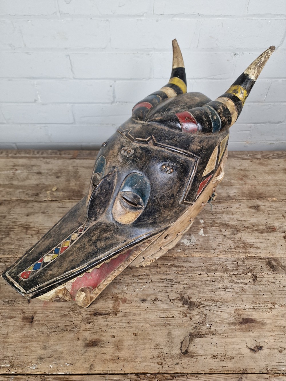 A carved wooden Guro Zamble helmet mask featuring an open-jawed snout with carved white teeth, red painted interior, dark horns, and woven fibre stitching along the base. View VII
