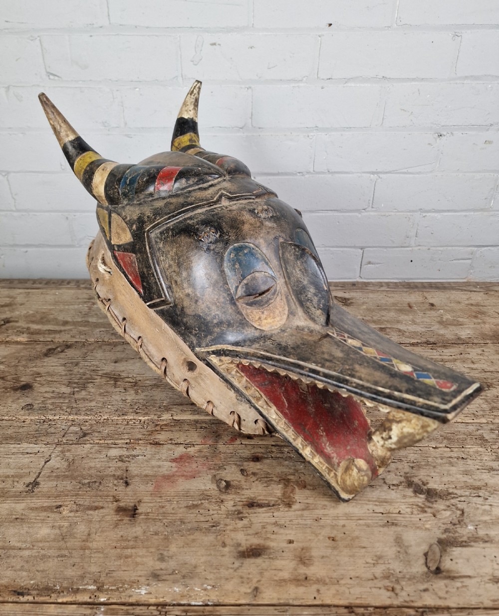 A carved wooden Guro Zamble helmet mask featuring an open-jawed snout with carved white teeth, red painted interior, dark horns, and woven fibre stitching along the base. View VIII