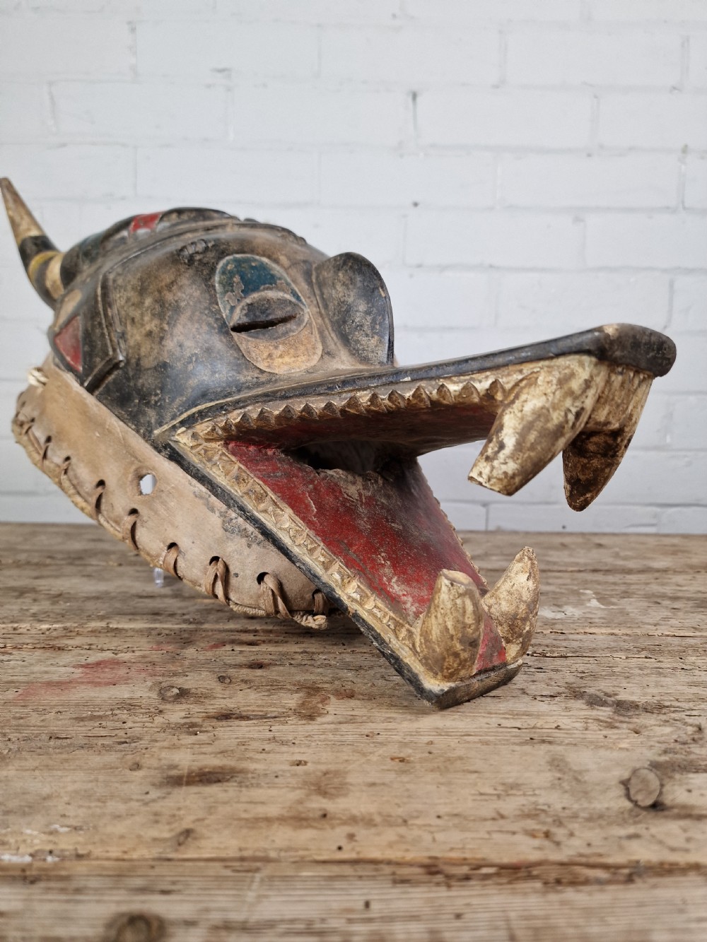 A carved wooden Guro Zamble helmet mask featuring an open-jawed snout with carved white teeth, red painted interior, dark horns, and woven fibre stitching along the base. View IX