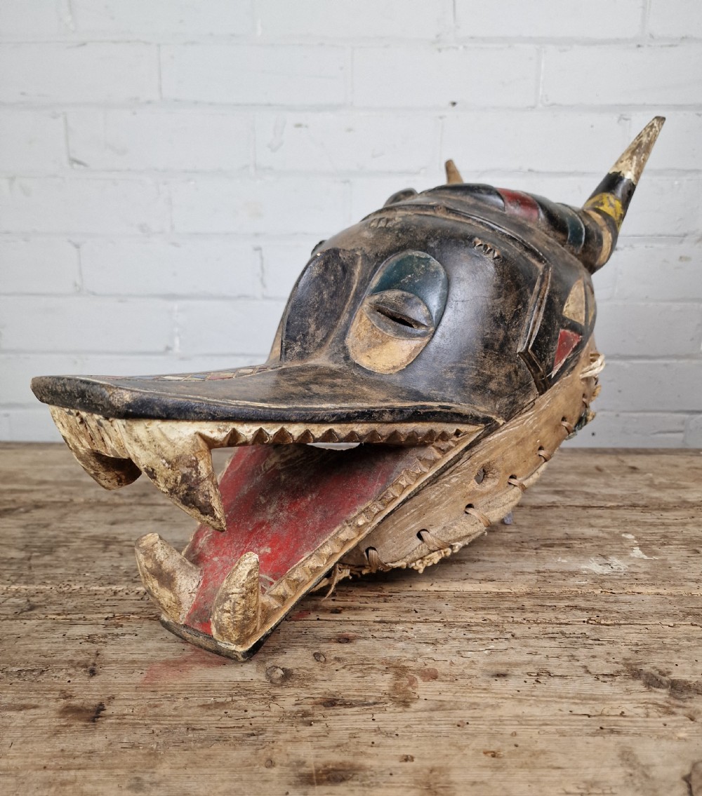 A carved wooden Guro Zamble helmet mask featuring an open-jawed snout with carved white teeth, red painted interior, dark horns, and woven fibre stitching along the base.