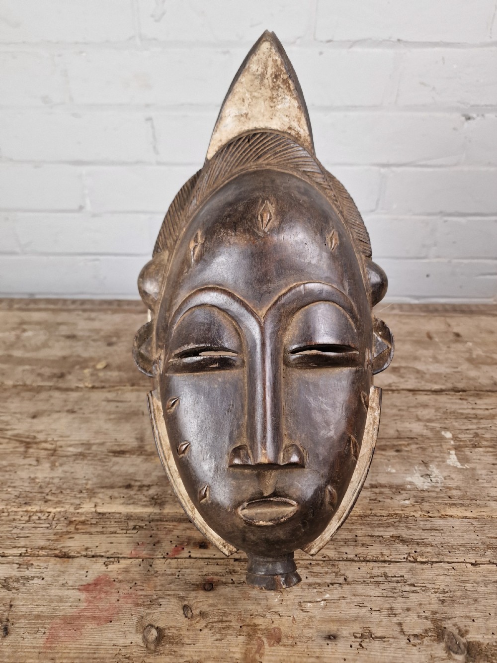 A carved dark wood Baule tribal mask featuring stylized facial features, facial scarification markings, a high crest hairstyle, and light pigment along the rim. View II