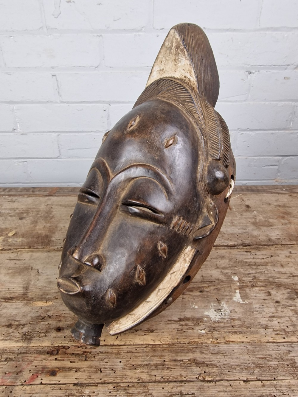 A carved dark wood Baule tribal mask featuring stylized facial features, facial scarification markings, a high crest hairstyle, and light pigment along the rim.