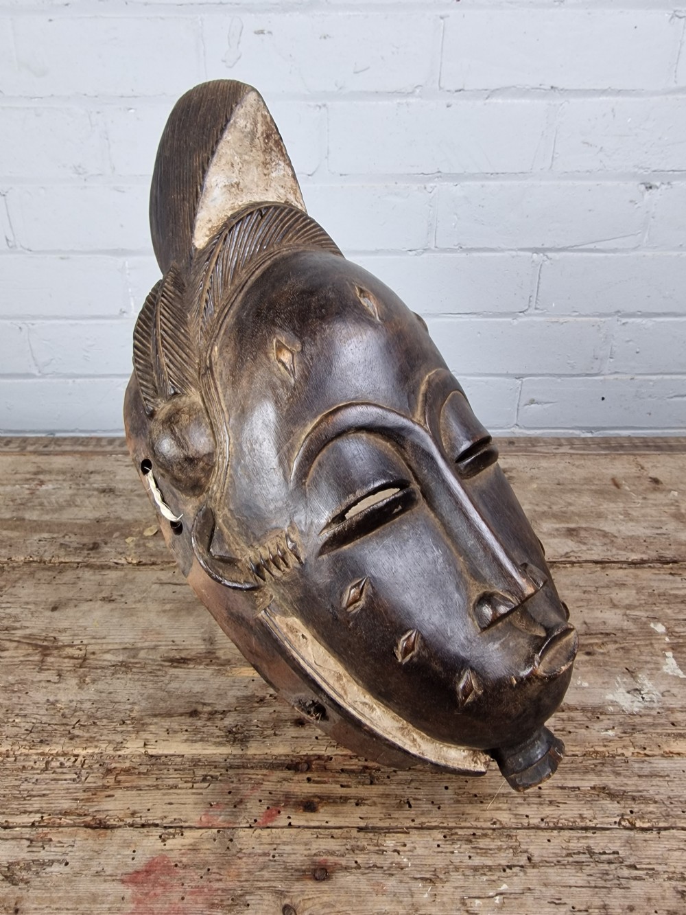 A carved dark wood Baule tribal mask featuring stylized facial features, facial scarification markings, a high crest hairstyle, and light pigment along the rim. View III