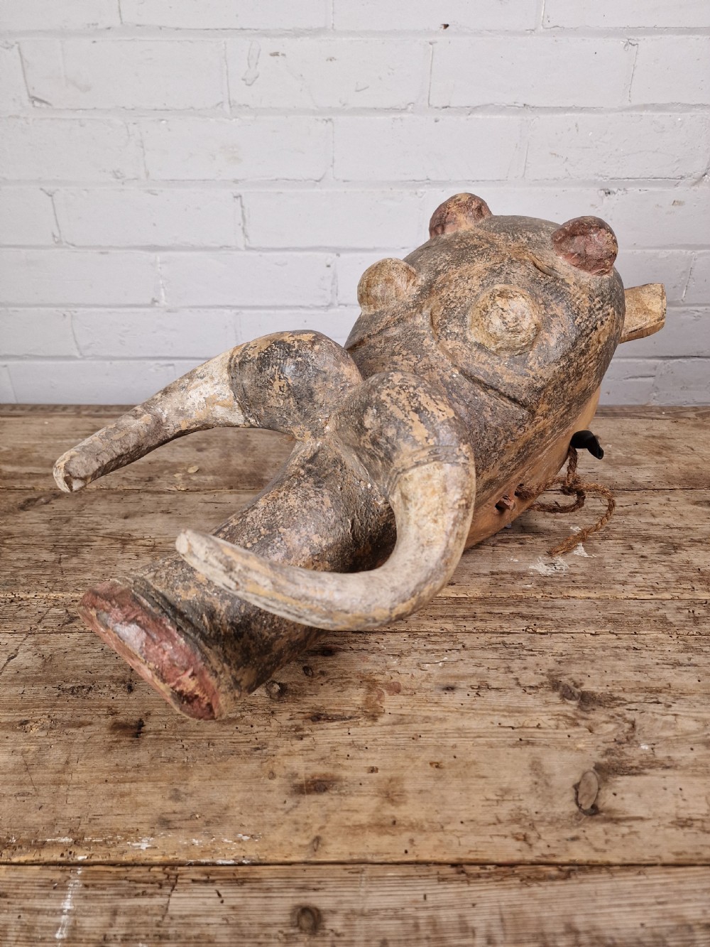 A hand-carved dark wood zoomorphic mask representing a warthog, featuring large curved tusks, rounded eye projections, and attached natural fibre ropes. View V