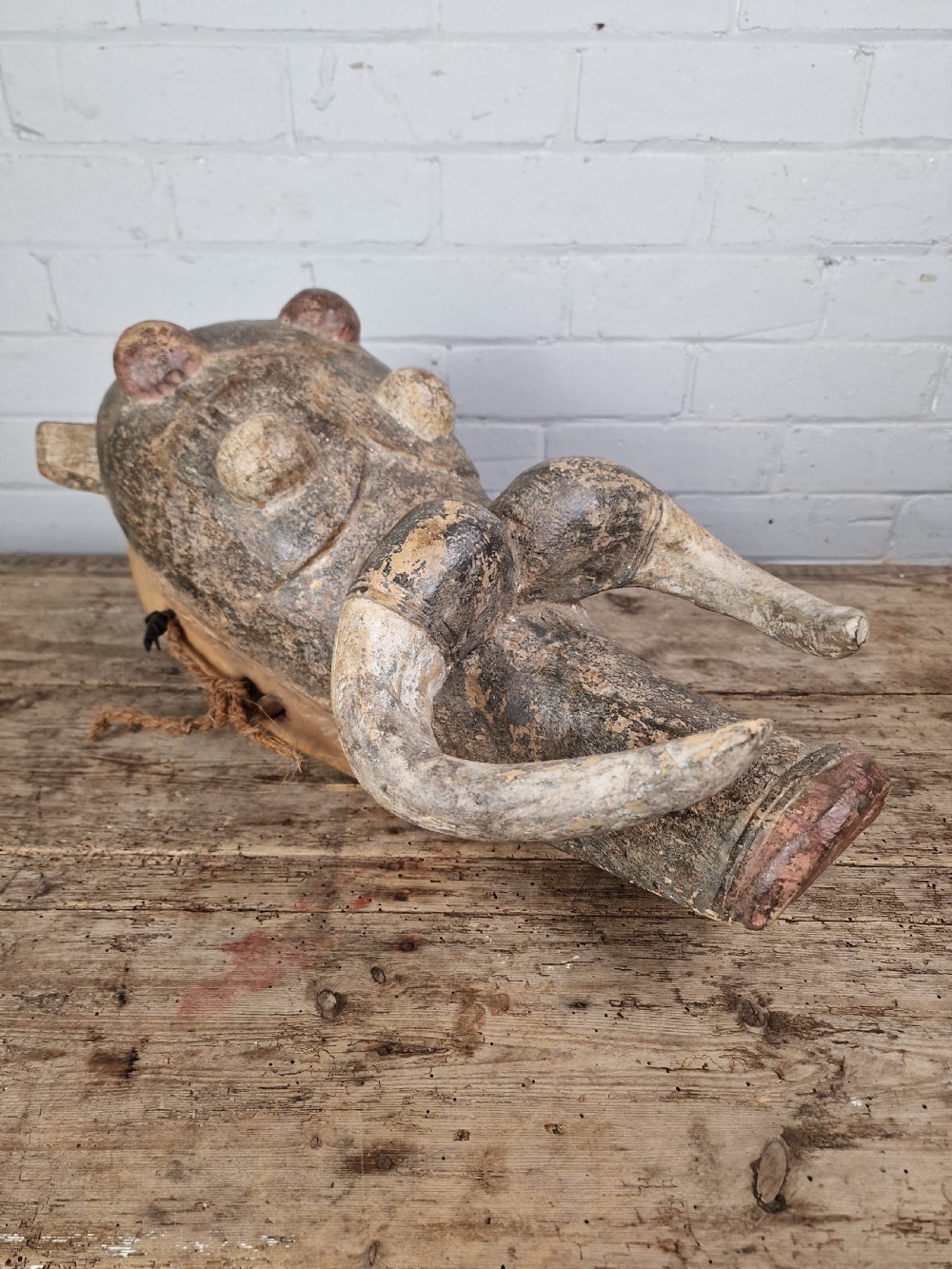 A hand-carved dark wood zoomorphic mask representing a warthog, featuring large curved tusks, rounded eye projections, and attached natural fibre ropes. View II