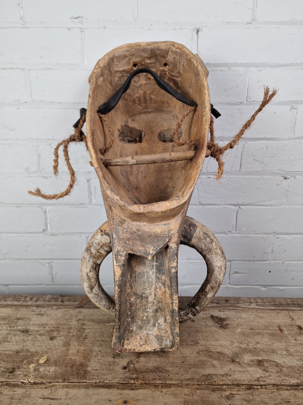 A hand-carved dark wood zoomorphic mask representing a warthog, featuring large curved tusks, rounded eye projections, and attached natural fibre ropes. View IV