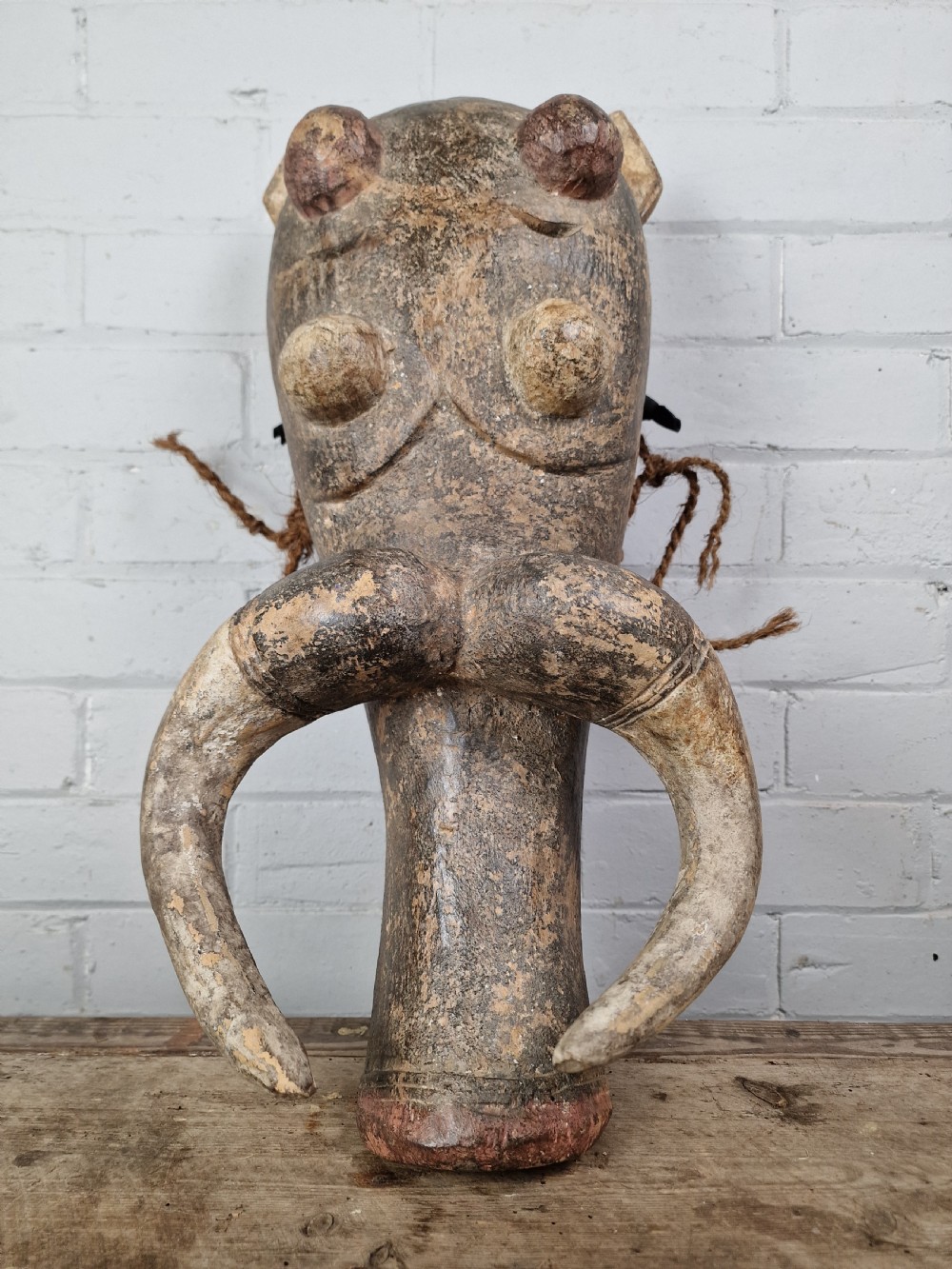 A hand-carved dark wood zoomorphic mask representing a warthog, featuring large curved tusks, rounded eye projections, and attached natural fibre ropes.