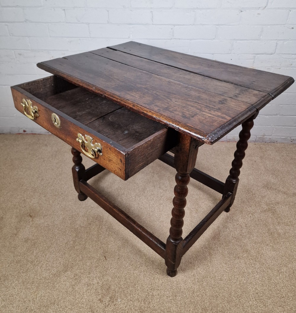 An 18th-century dark oak side table featuring four bobbin-turned legs, a square top, a single drawer with brass handles, and a box stretcher. View VI