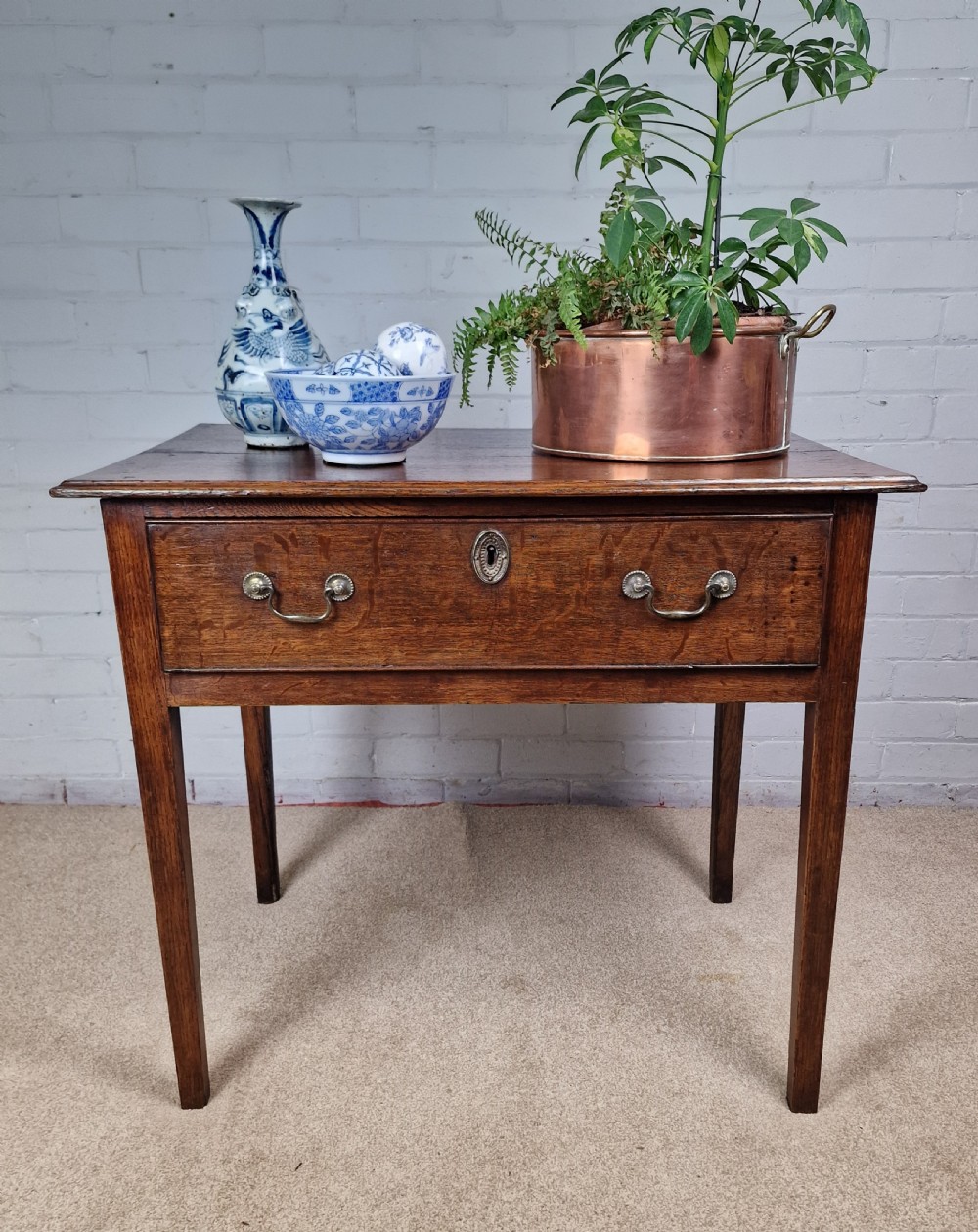 An 18th-century antique oak lowboy side table featuring a single wide drawer with brass handles and square tapered legs.