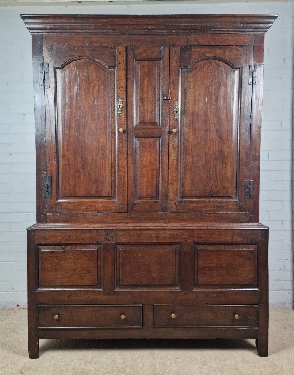 A tall Georgian dark oak livery cupboard featuring arched panel doors, iron hinges, three lower panels, and two drawers at the base.