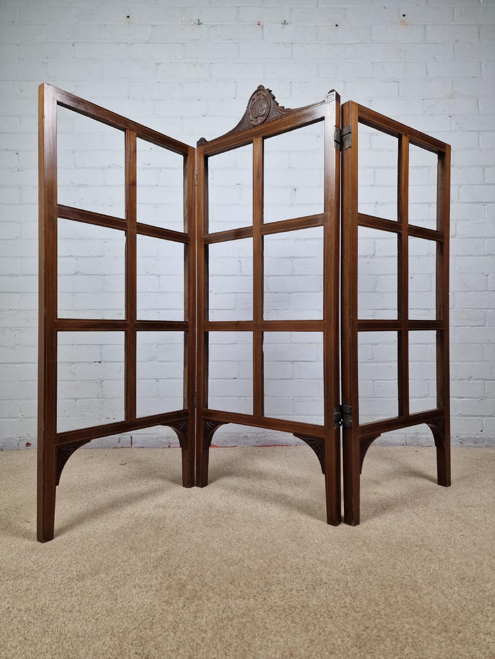 An Edwardian three-panel mahogany folding screen featuring a central carved crest, glazed grid panes, and bracket feet. View IV