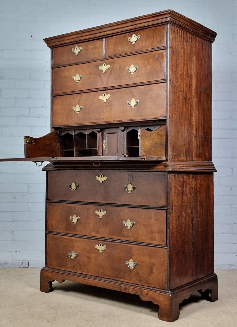 An early eighteenth-century Georgian walnut secretaire chest on chest featuring brass drop handles, a drop-front desk compartment with fitted pigeonholes, and bracket feet.