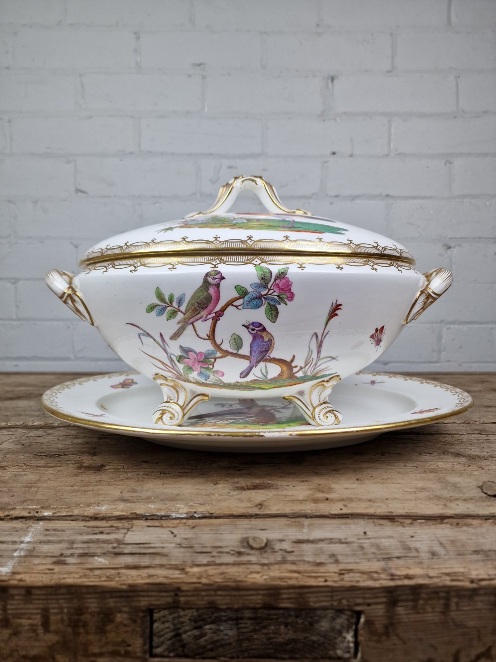 An antique white porcelain soup tureen featuring hand-painted bird motifs, gilded scroll handles, four scroll feet, and a matching underplate.