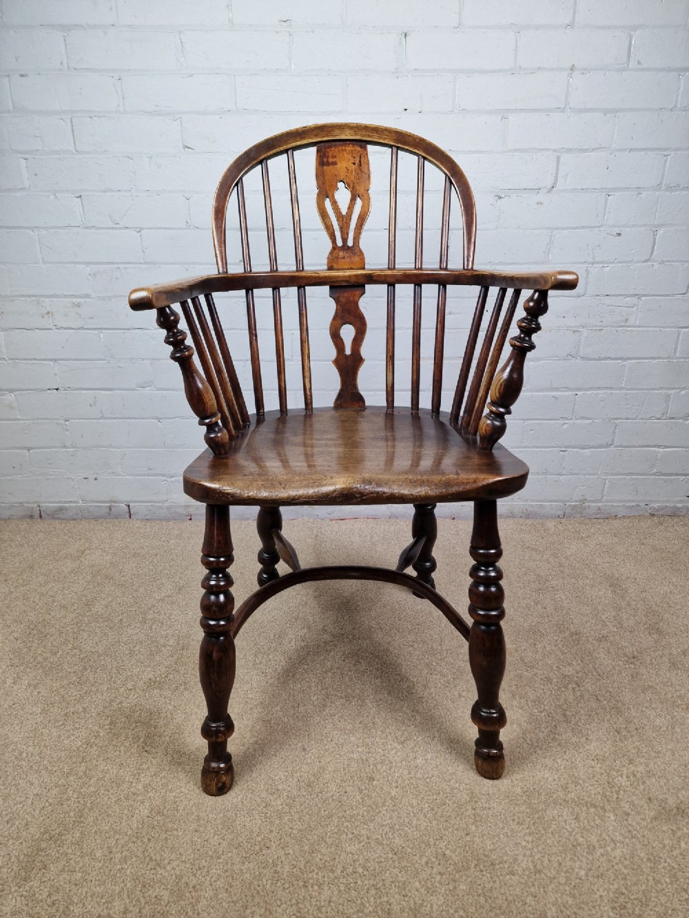 A 19th-century low-back wooden Windsor armchair featuring a curved top rail, pierced central splat, vertical spindles, turned legs, and a crinoline stretcher.