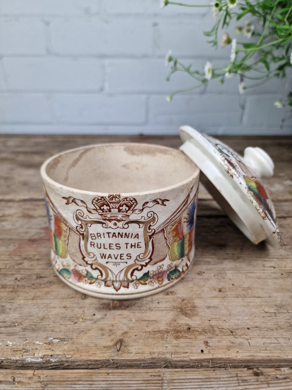 A cylindrical cream ceramic butter dish with a lid, decorated with a brown transfer print featuring flags, a crown, and the inscription Britannia Rules The Waves. View II