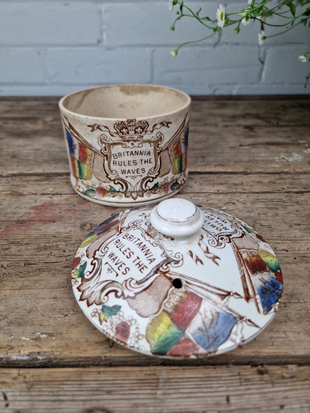A cylindrical cream ceramic butter dish with a lid, decorated with a brown transfer print featuring flags, a crown, and the inscription Britannia Rules The Waves. View III