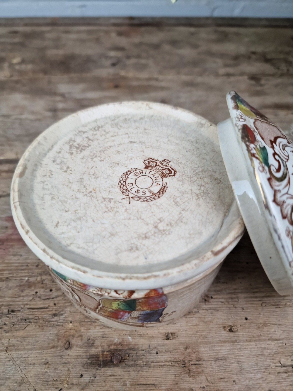 A cylindrical cream ceramic butter dish with a lid, decorated with a brown transfer print featuring flags, a crown, and the inscription Britannia Rules The Waves. View IV