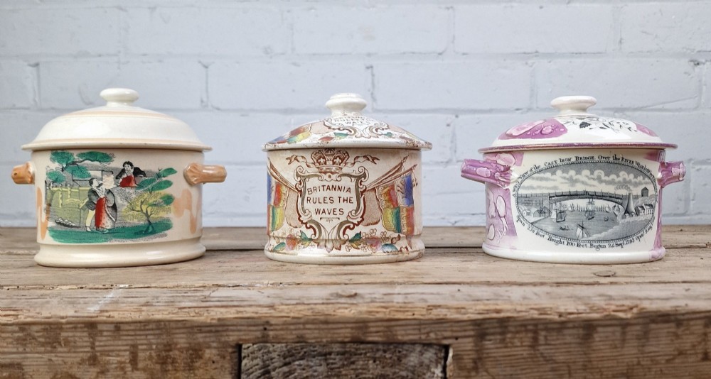 A cylindrical cream ceramic butter dish with a lid, decorated with a brown transfer print featuring flags, a crown, and the inscription Britannia Rules The Waves. View V