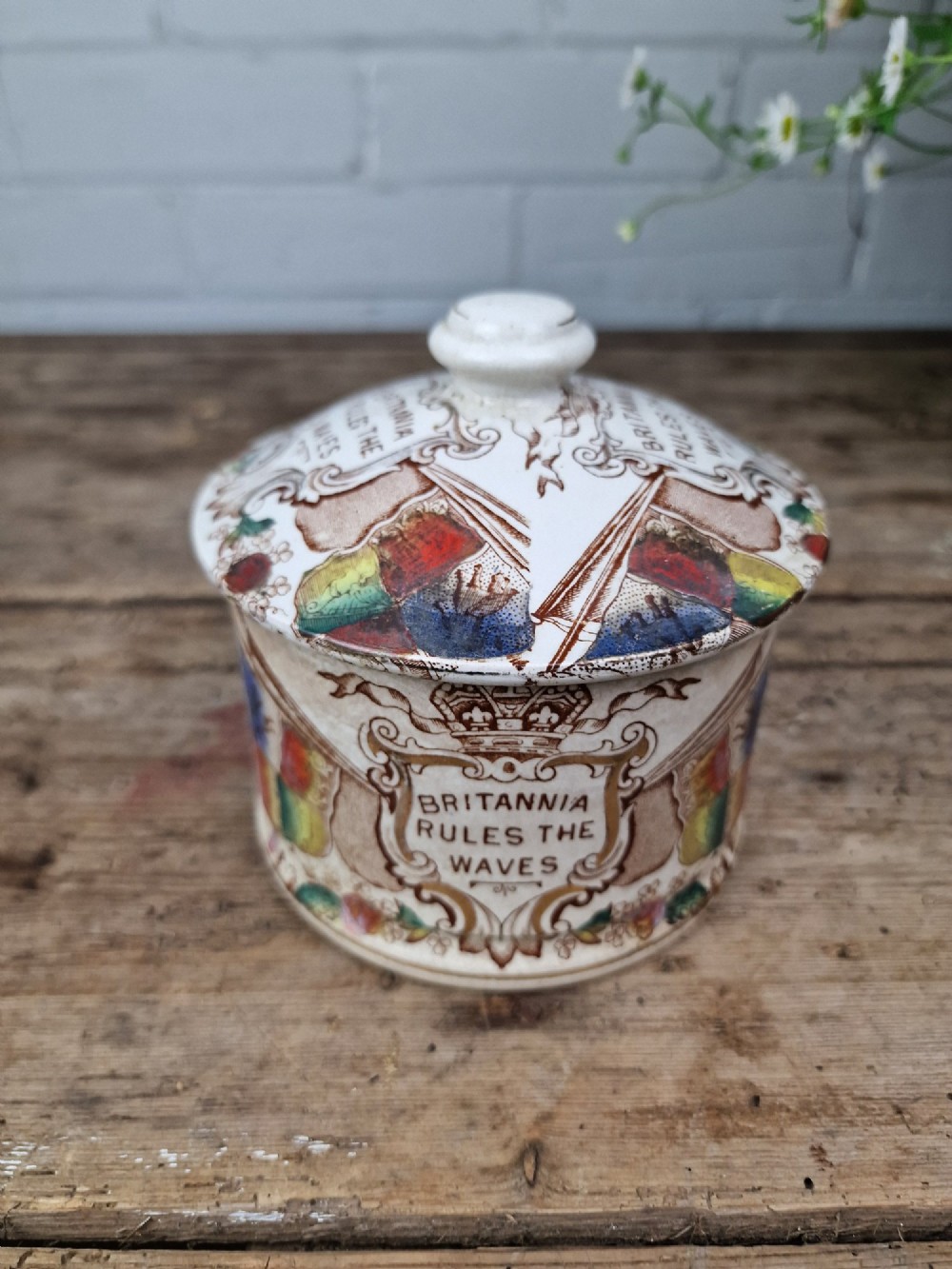 A cylindrical cream ceramic butter dish with a lid, decorated with a brown transfer print featuring flags, a crown, and the inscription Britannia Rules The Waves. View VI