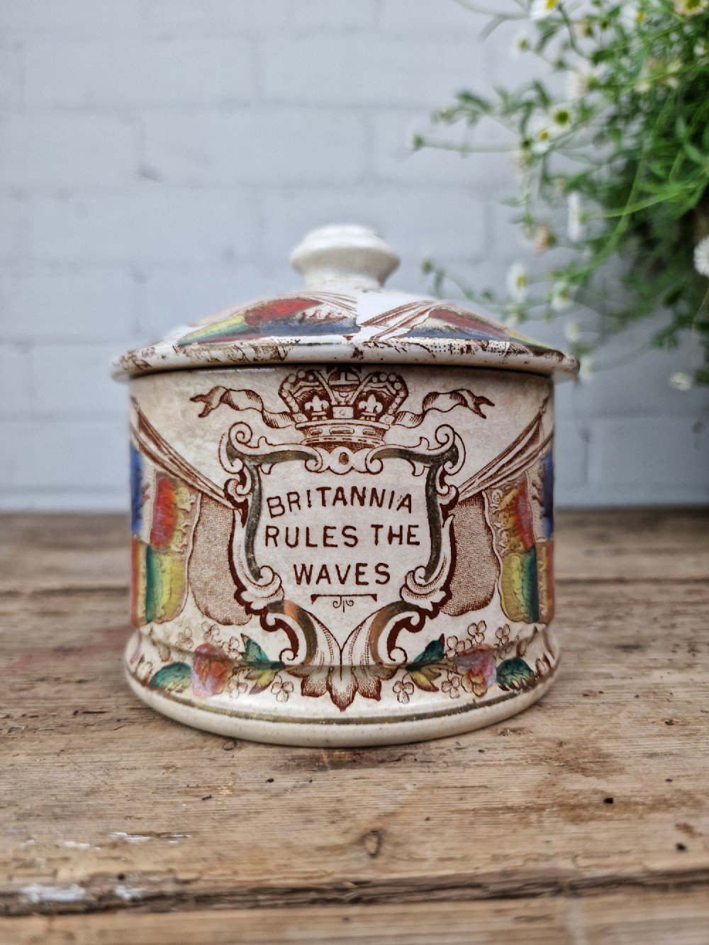 A cylindrical cream ceramic butter dish with a lid, decorated with a brown transfer print featuring flags, a crown, and the inscription Britannia Rules The Waves.