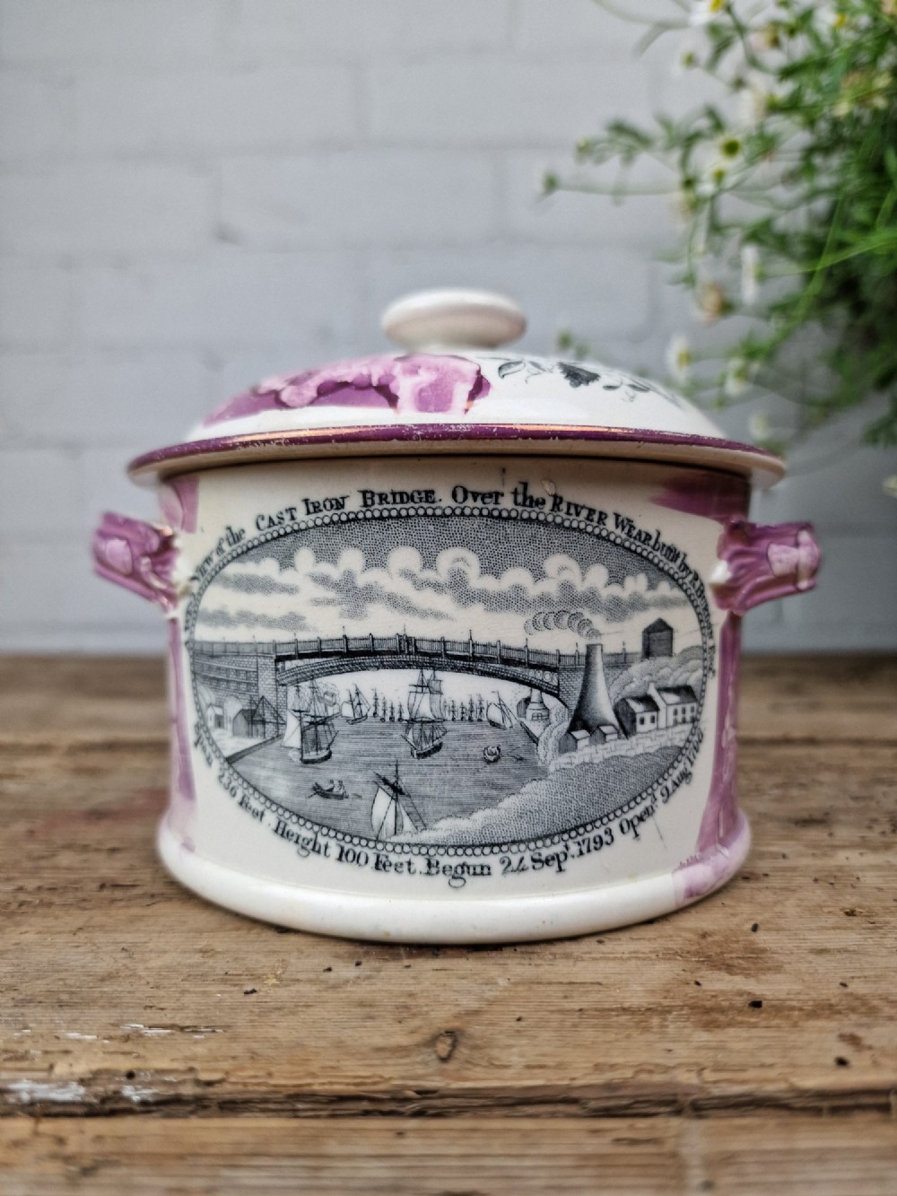 A cream earthenware butter dish with pink splash lustre accents, side handles, and a black transfer print depicting a bridge over a river.