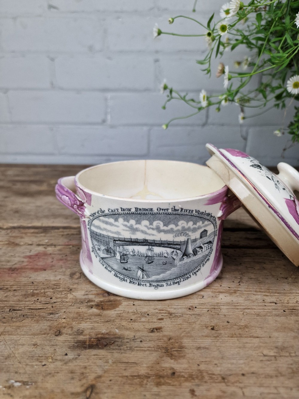 A cream earthenware butter dish with pink splash lustre accents, side handles, and a black transfer print depicting a bridge over a river. View III
