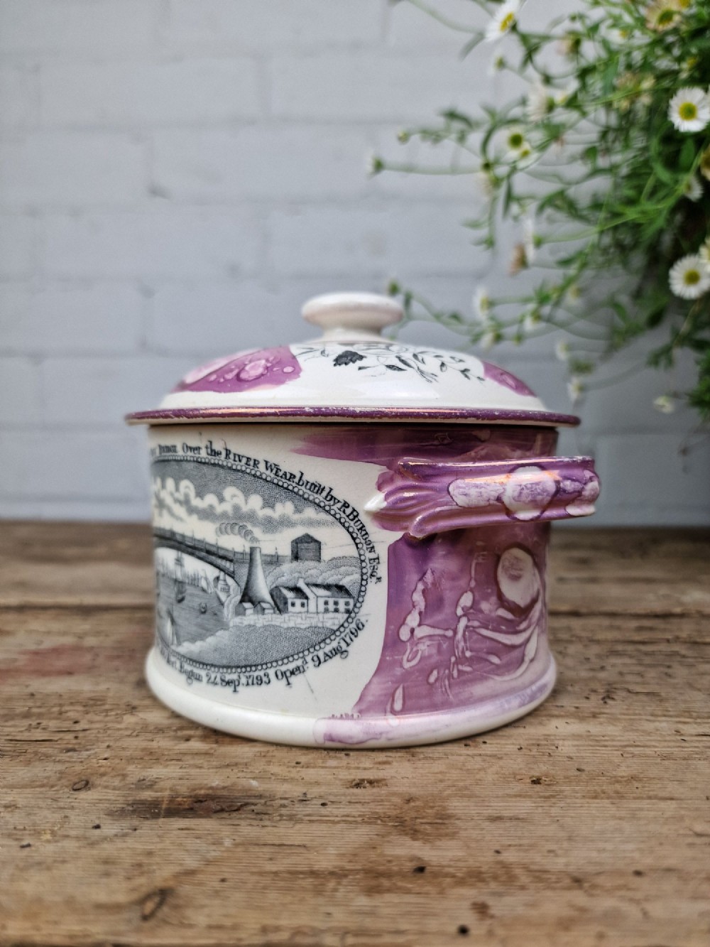 A cream earthenware butter dish with pink splash lustre accents, side handles, and a black transfer print depicting a bridge over a river. View IV