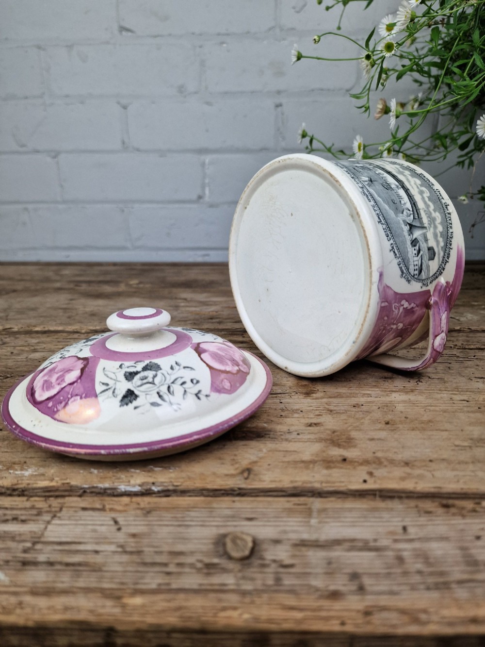 A cream earthenware butter dish with pink splash lustre accents, side handles, and a black transfer print depicting a bridge over a river. View V