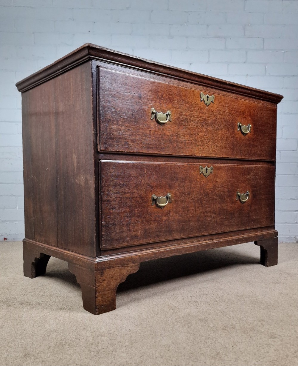 An 18th-century dark oak chest of drawers featuring two wide drawers with brass handles, standing on shaped bracket feet. View IV