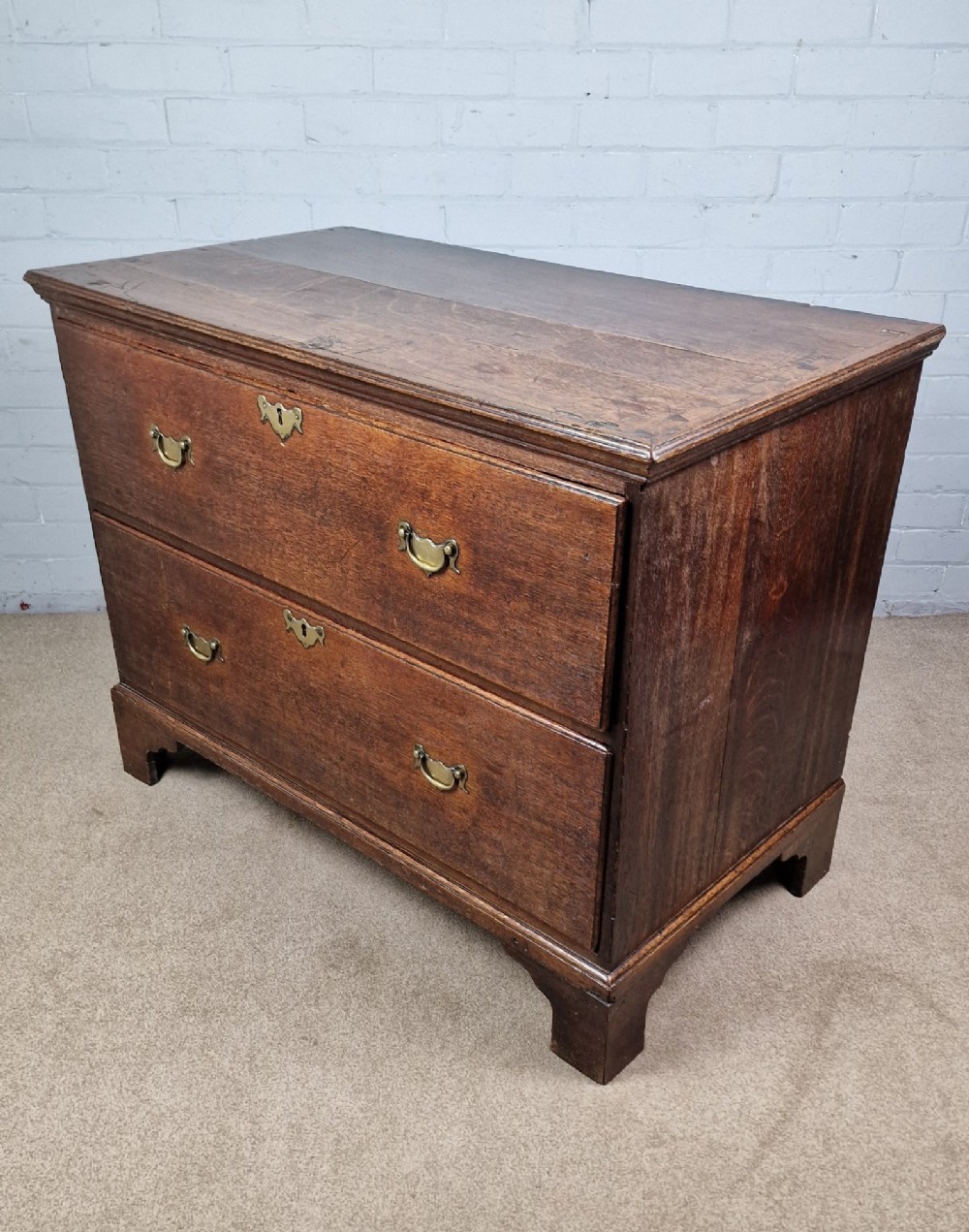 An 18th-century dark oak chest of drawers featuring two wide drawers with brass handles, standing on shaped bracket feet. View V