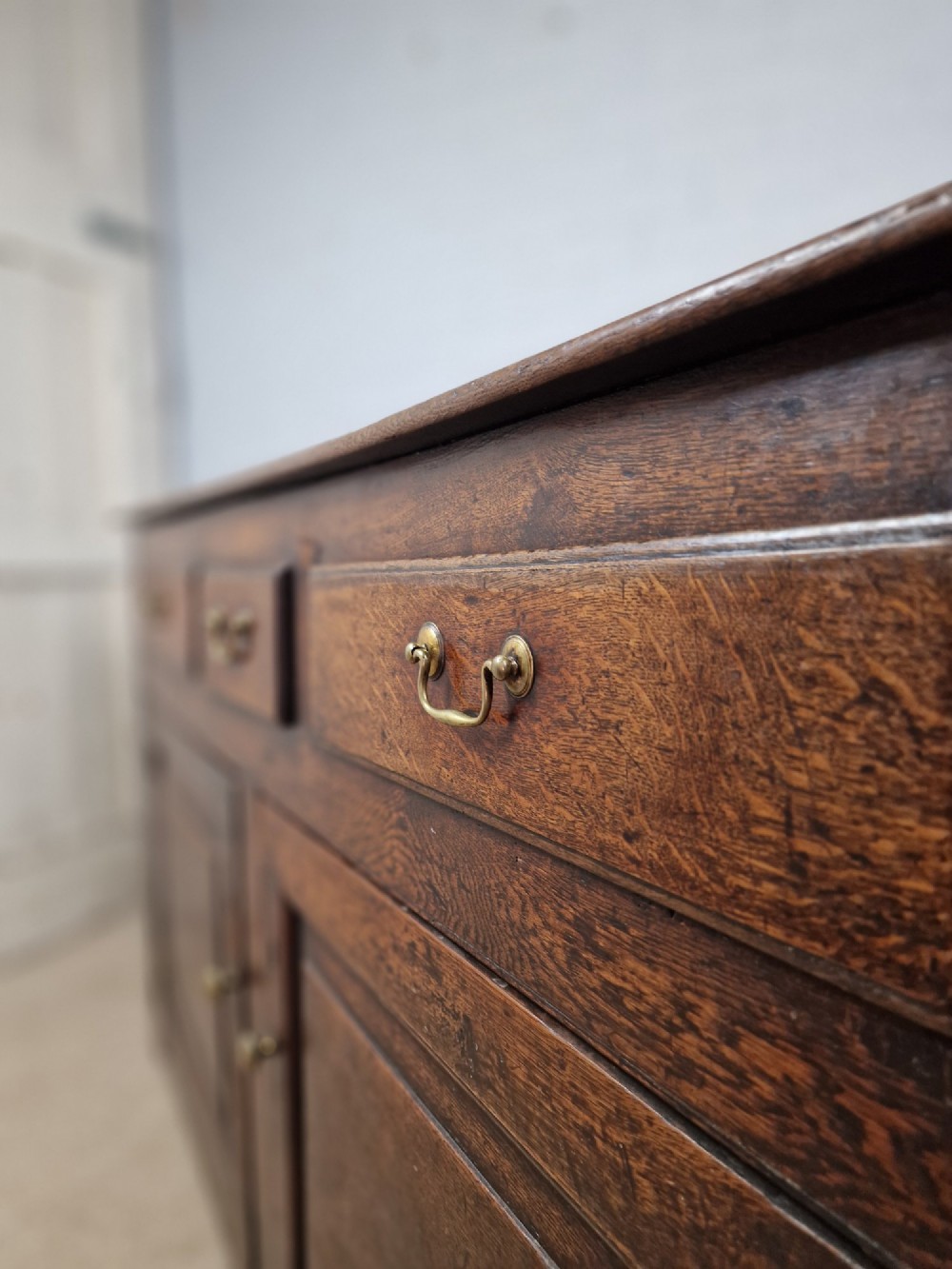 An antique dark oak dresser base featuring three upper drawers above two fielded panel cupboard doors with brass drop handles and a stepped upper gallery. View II