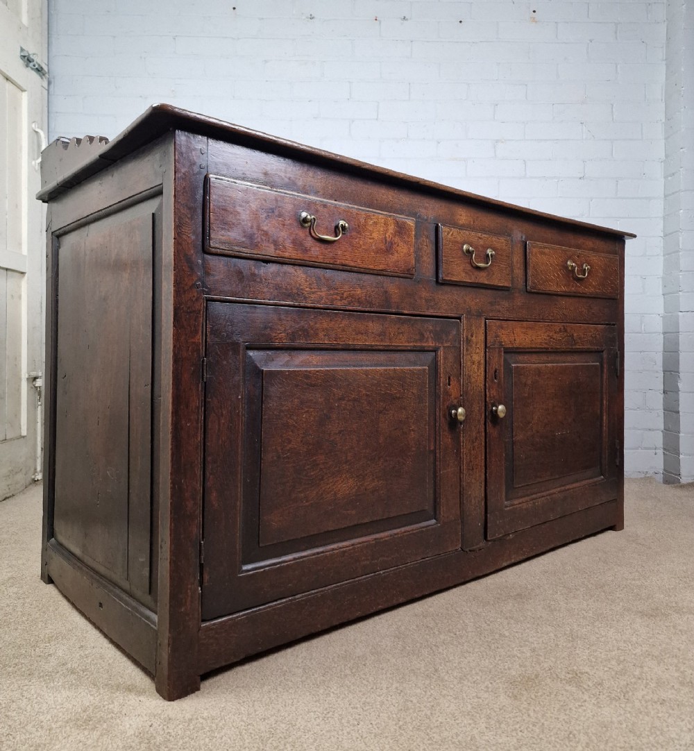 An antique dark oak dresser base featuring three upper drawers above two fielded panel cupboard doors with brass drop handles and a stepped upper gallery. View IV