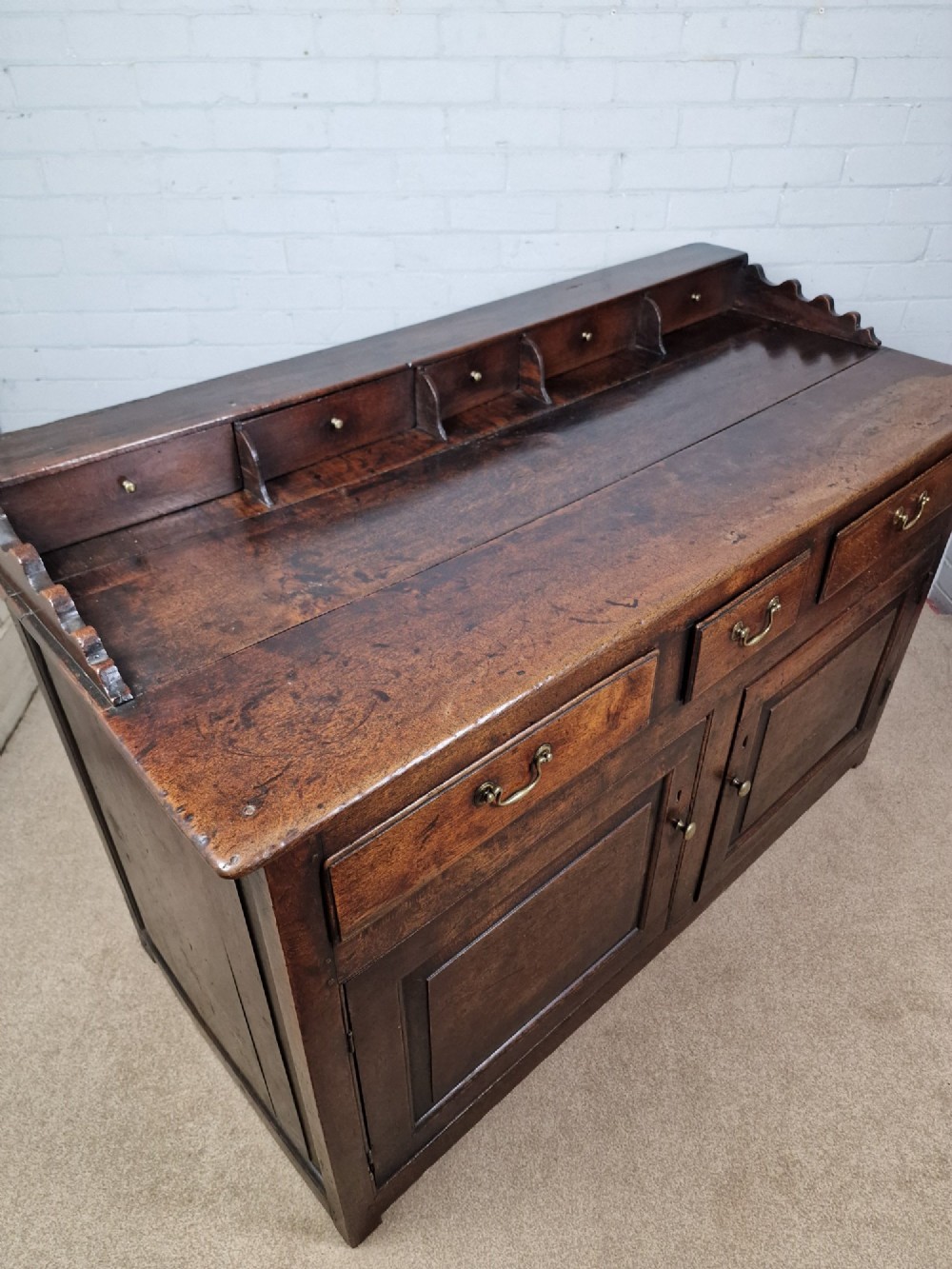 An antique dark oak dresser base featuring three upper drawers above two fielded panel cupboard doors with brass drop handles and a stepped upper gallery. View V
