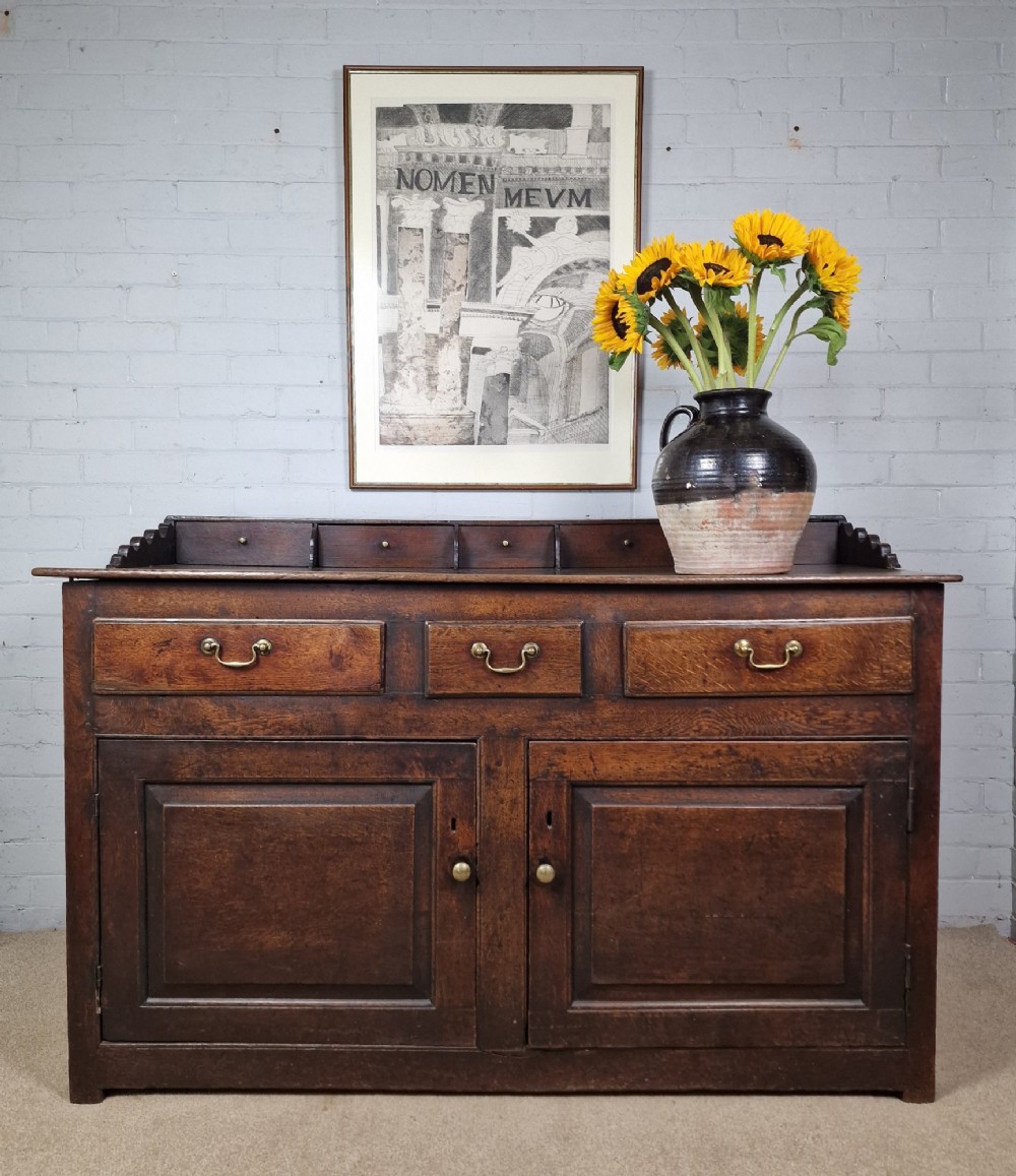 An antique dark oak dresser base featuring three upper drawers above two fielded panel cupboard doors with brass drop handles and a stepped upper gallery.